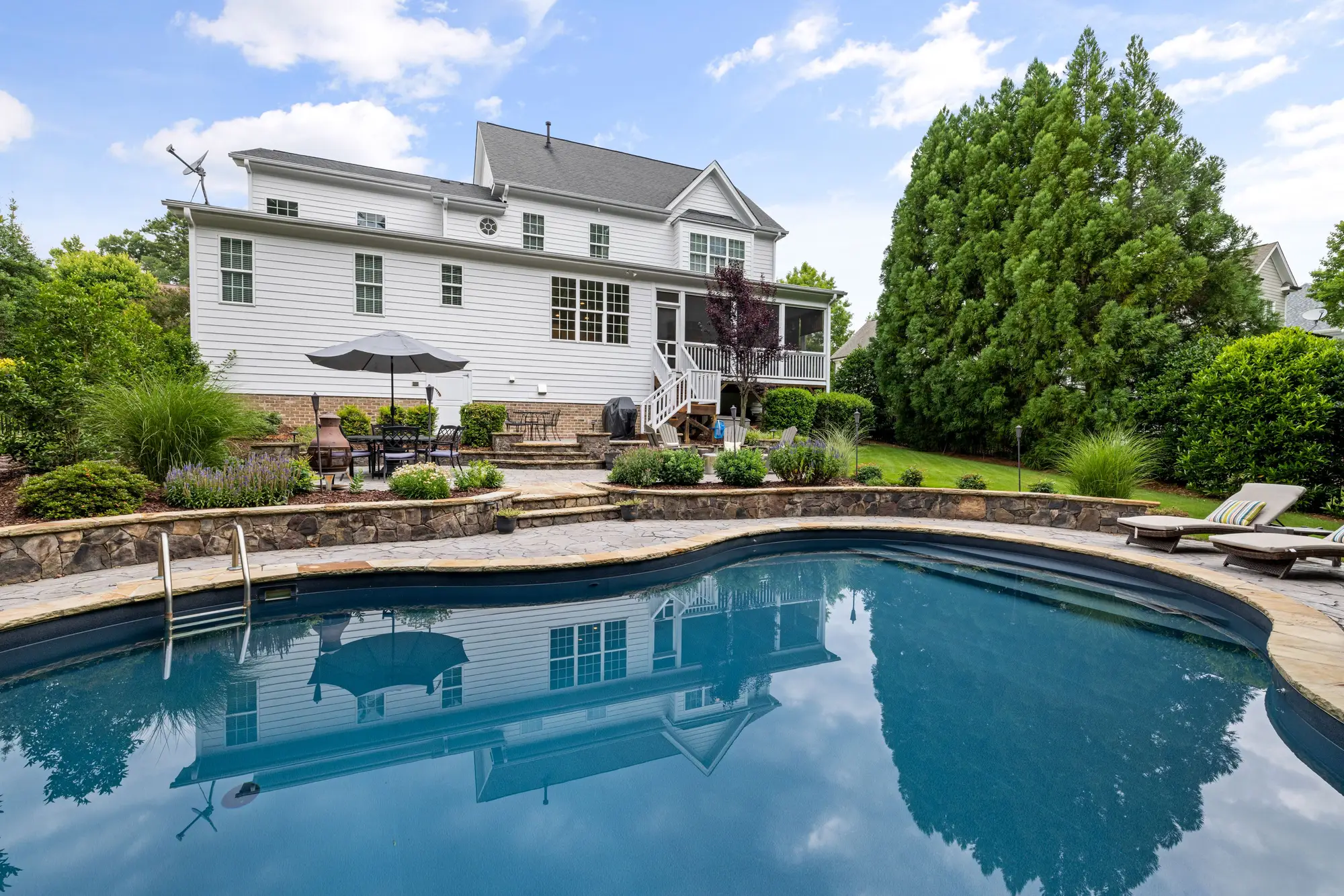 The back of a two-storey clapboard house seen across an in-ground pool, with a patio, deck stairs and a lawn running to tall trees