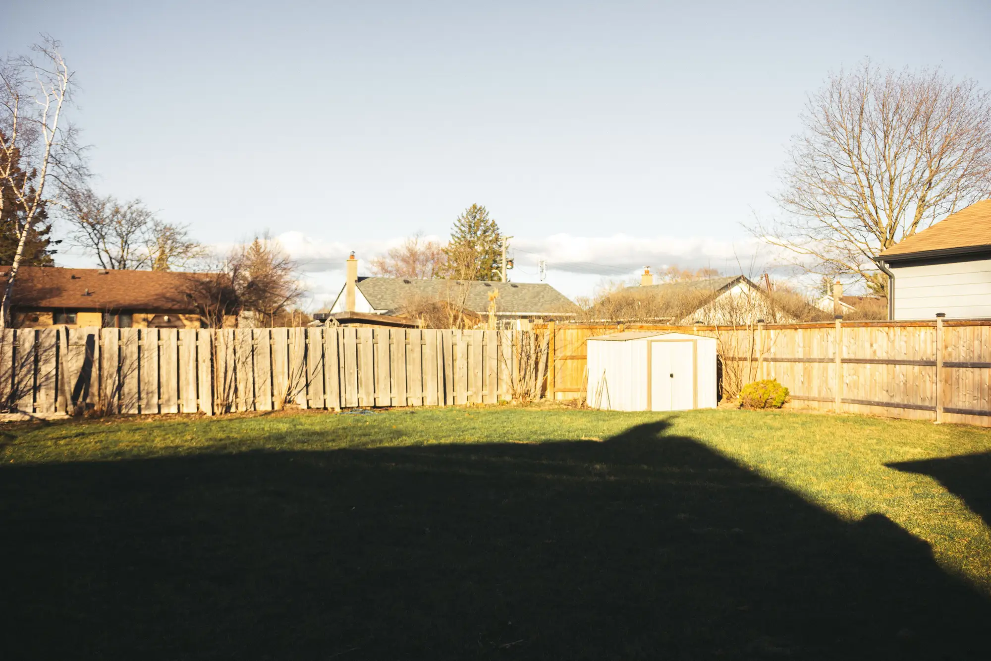 A small metal shed at the back of a fenced suburban lawn, with neighbouring roofs beyond the fence